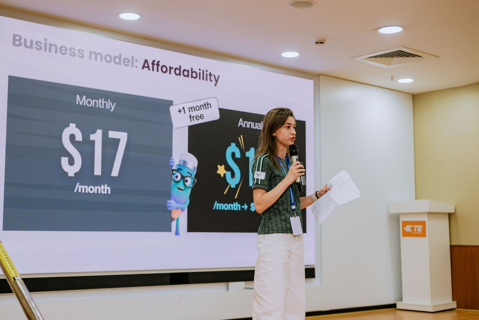 O membră a echipei prezintă pe scenă slide-ul „Business model: Affordability”, cu prețul de $17 pe lună și mascota Tuto