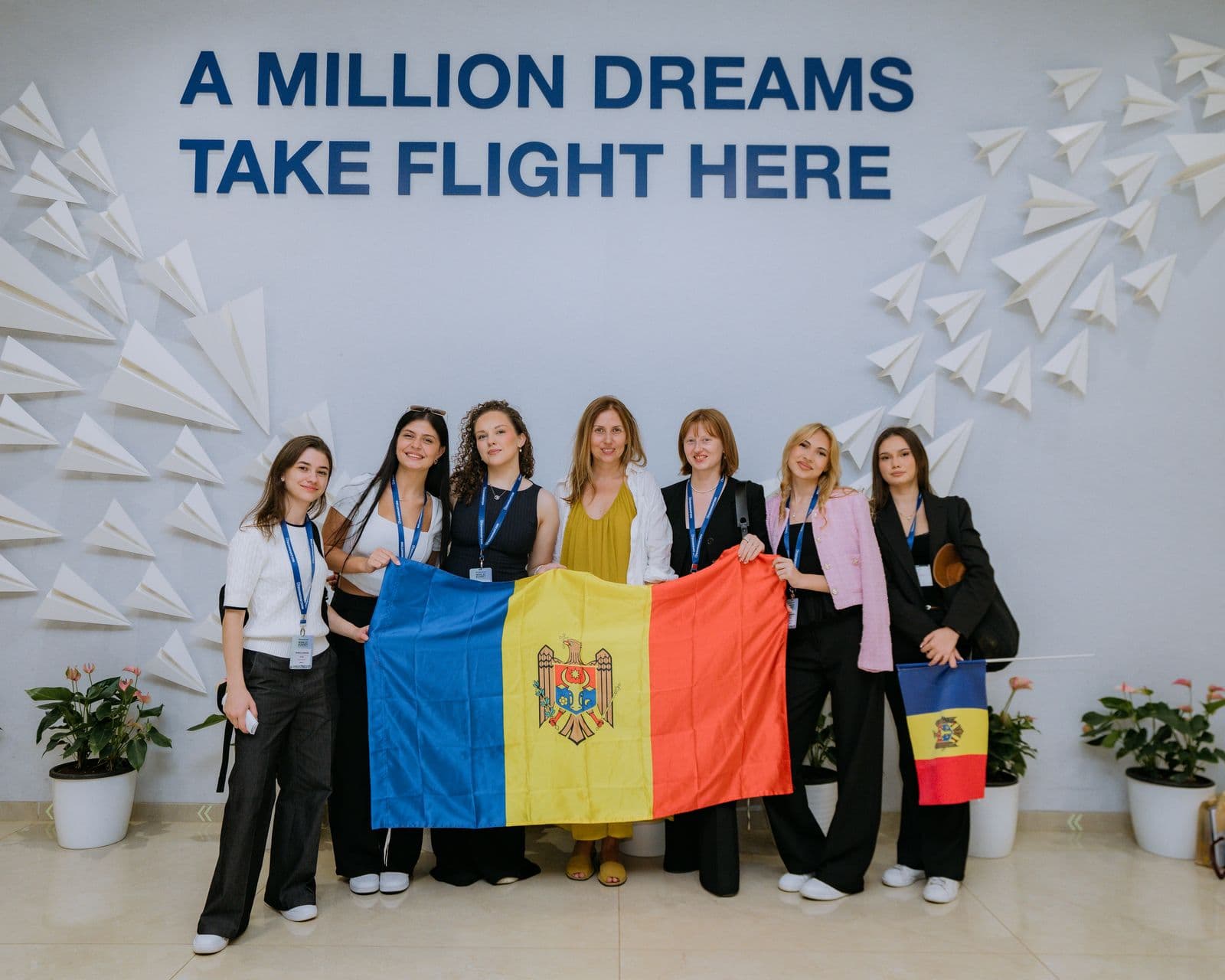 Echipa și însoțitorii cu un steag mare al Moldovei, în fața peretelui cu textul A Million Dreams Take Flight Here